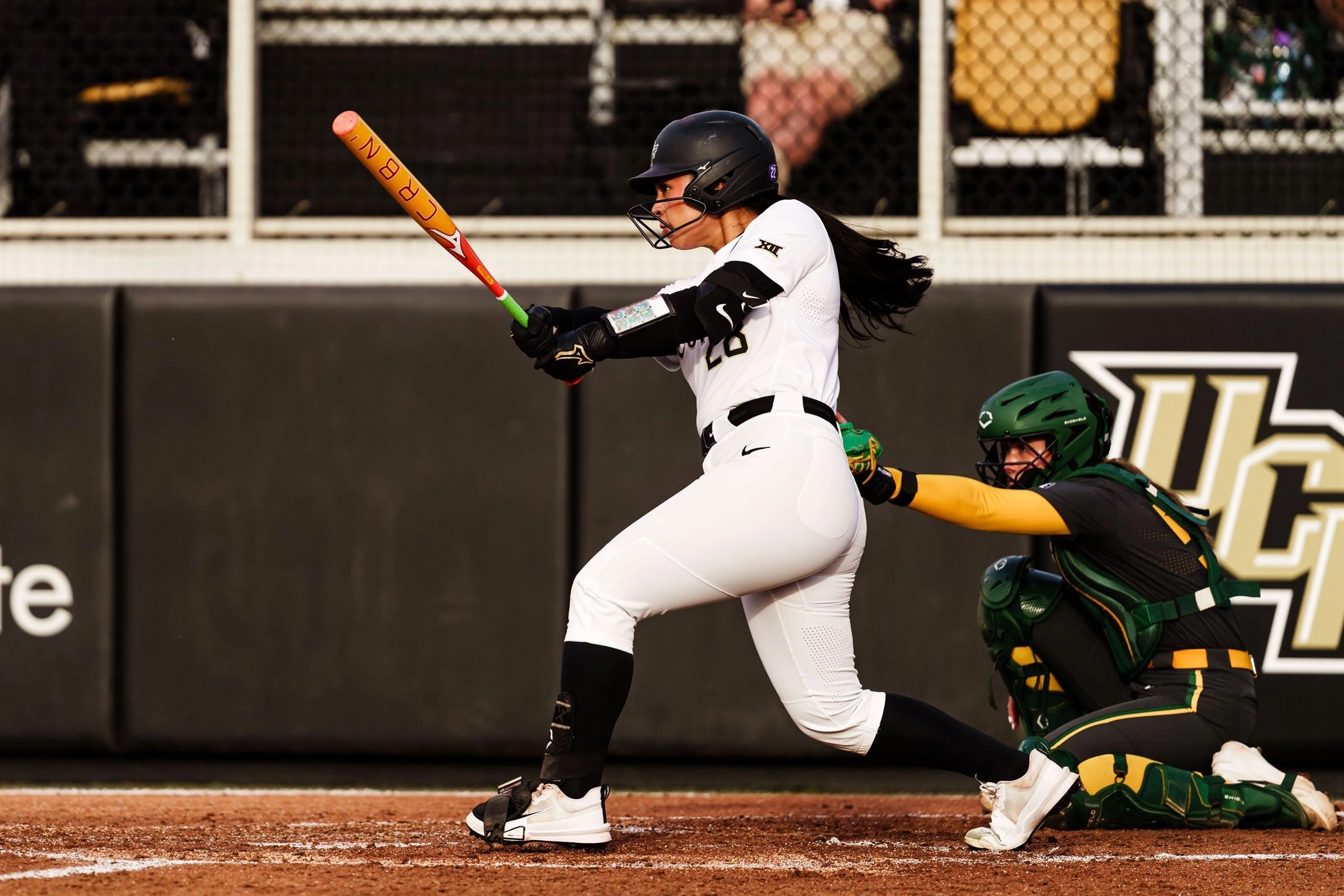 Izzy Mertes following through on a swing in UCF's white home uniform as the catcher looks on
